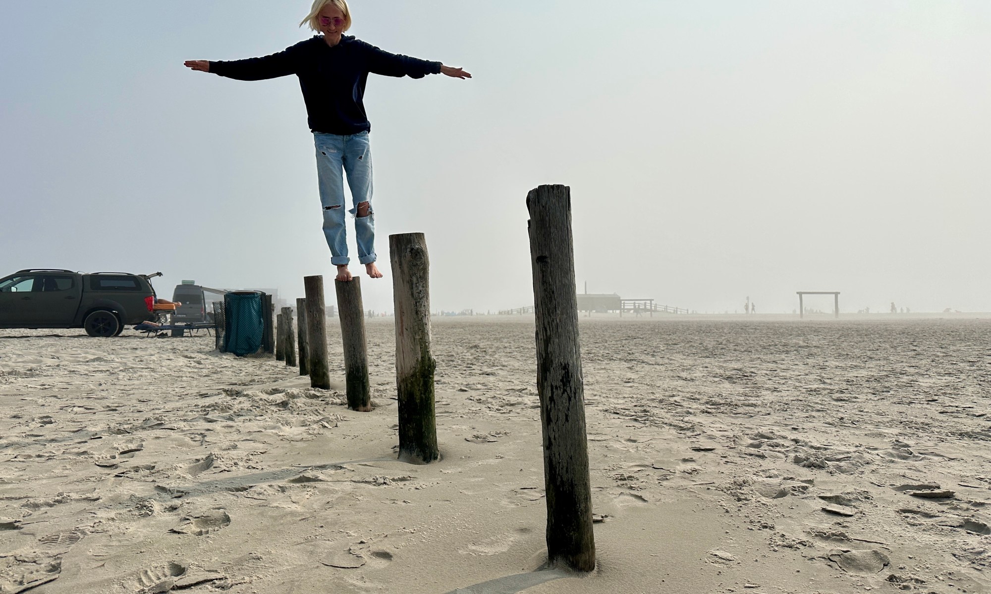 Frau steht barfuß auf Holzpfählen am Strand von Sankt Peter-Ording – Symbol für innere Stärke, Balance und Orientierung auf unsicherem Untergrund.